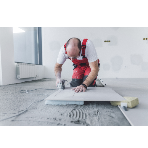 Worker wearing protective goggles and red overalls using an electric tile cutter to cut a large ceramic tile on a floor under construction, demonstrating precise outillage technique.
